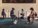 Vier jongeren met een VR bril en koptelefoon zitten voor een schilderij